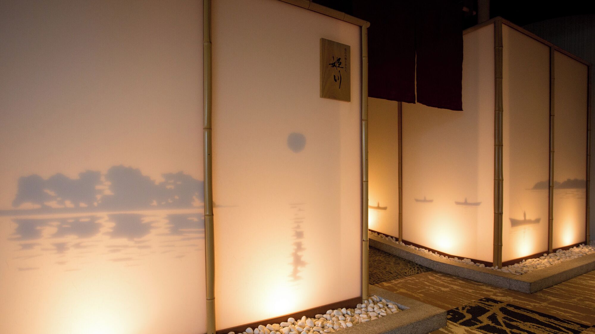 A row of traditional Japanese-style buildings with illuminated interiors, a calm water body in the background, and a clear night sky.