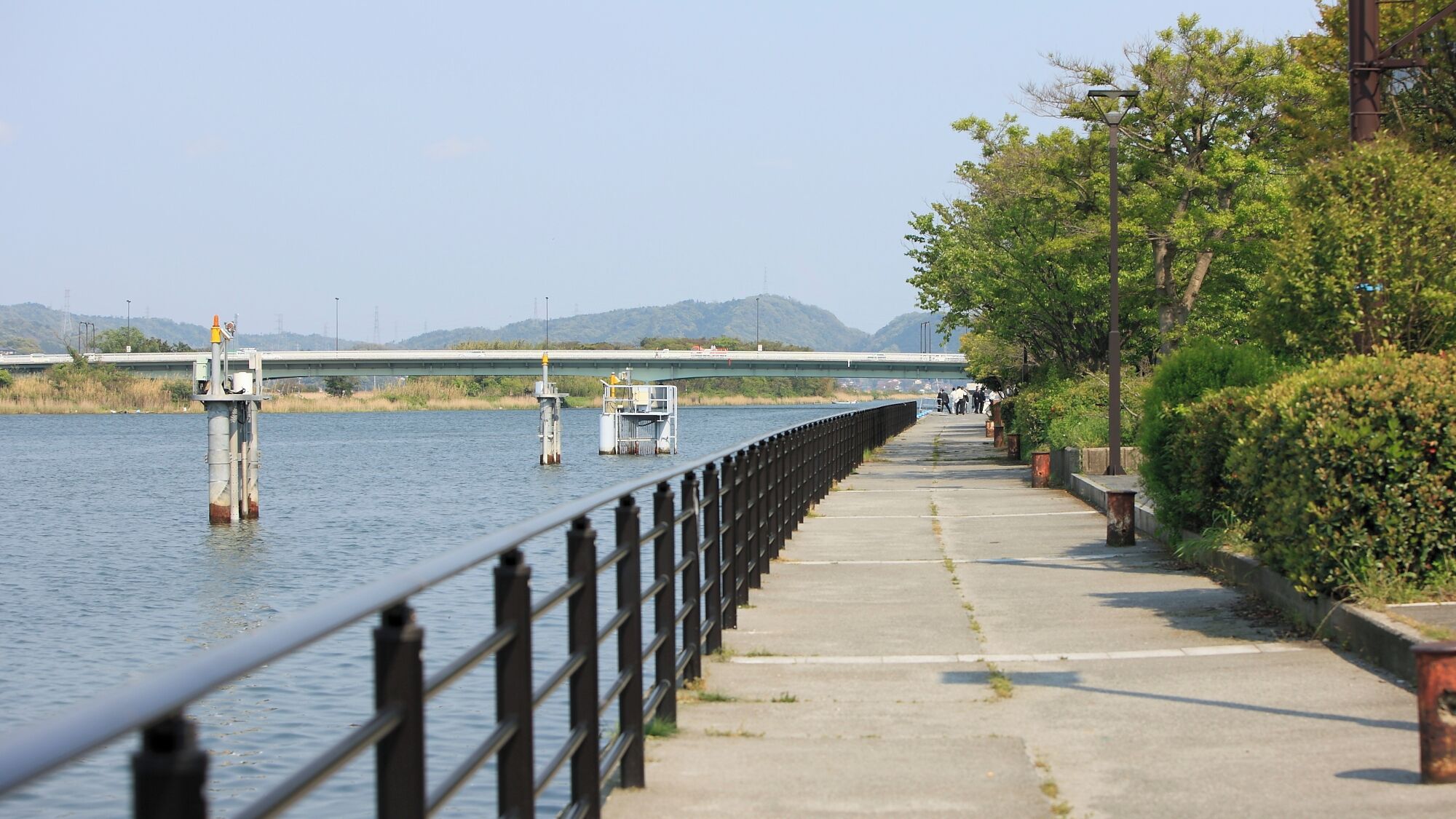 A riverside walkway with a metal railing, a concrete path, and a bridge in the background.