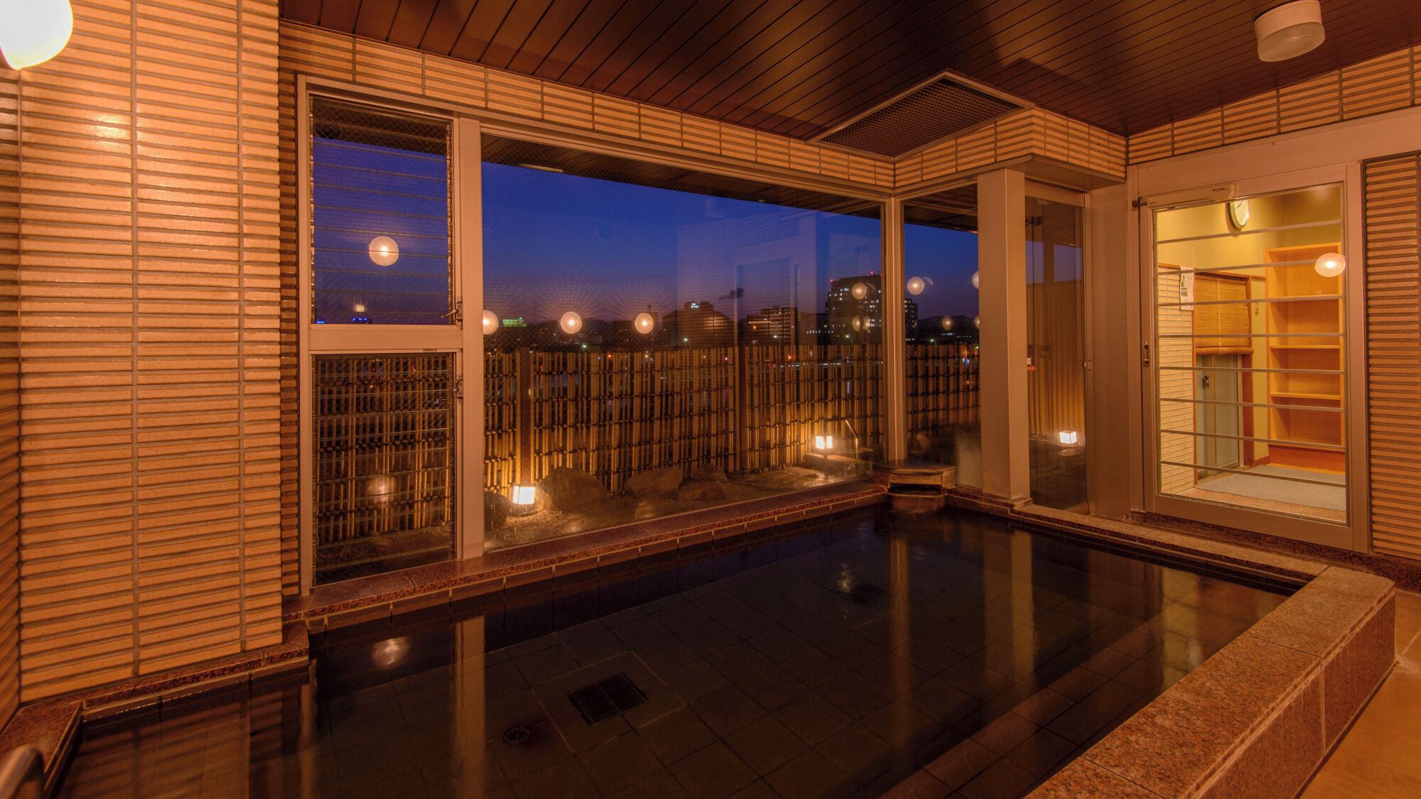 A modern indoor pool area with large windows, a bamboo fence, and a view of the cityscape at dusk.