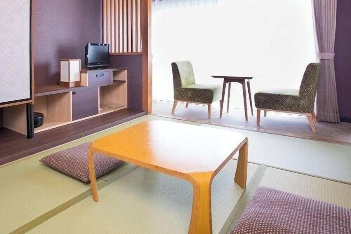 Una stanza con pavimento in tatami, un tavolino basso, due sedie, un televisore e un'ampia finestra con tende.