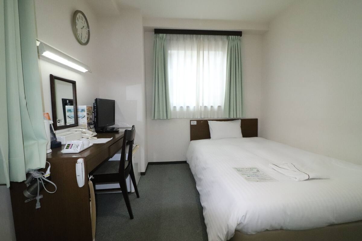 Habitación de hotel con cama, escritorio con computadora, reloj y ventana con cortinas.