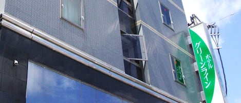 Un edificio de varios pisos con grandes ventanales que reflejan nubes, un letrero verde con caracteres japoneses y un escape de incendios visible.