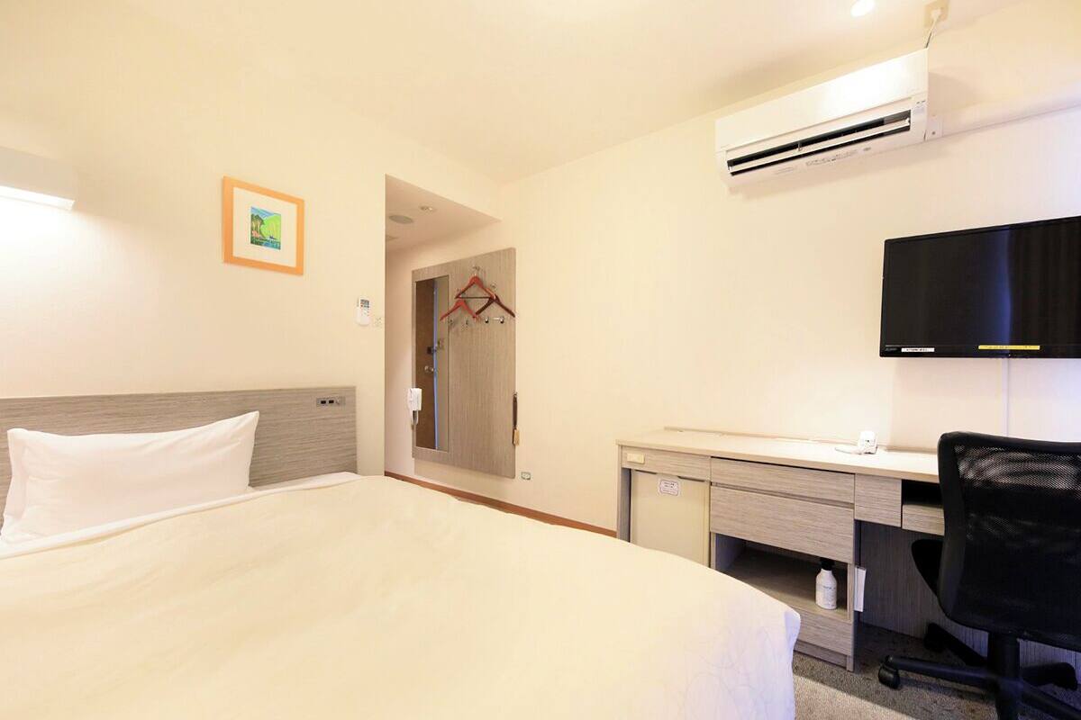 A hotel room with a bed, a desk, a chair, a TV, and an air conditioning unit.