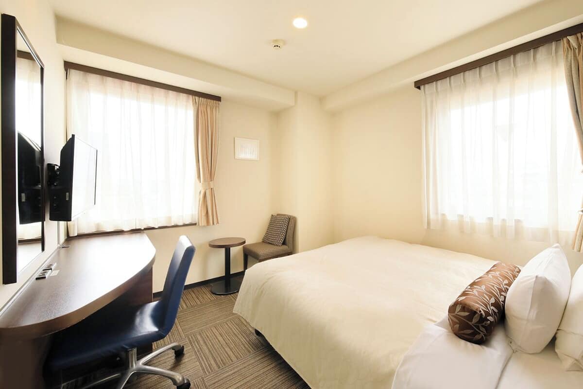 A hotel room with a bed, a desk with a chair, a small table, a chair, and a television.