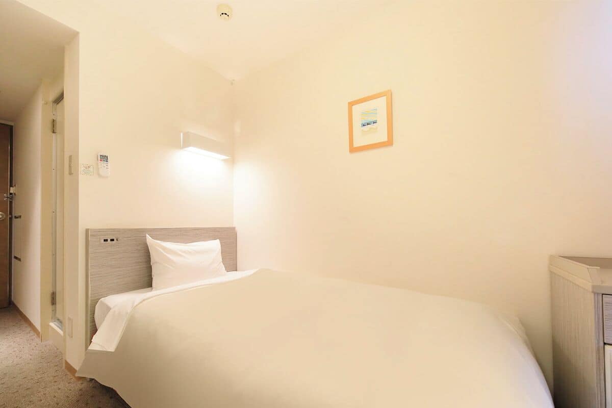 A hotel room with a bed, a nightstand, a wall-mounted light, and a door.