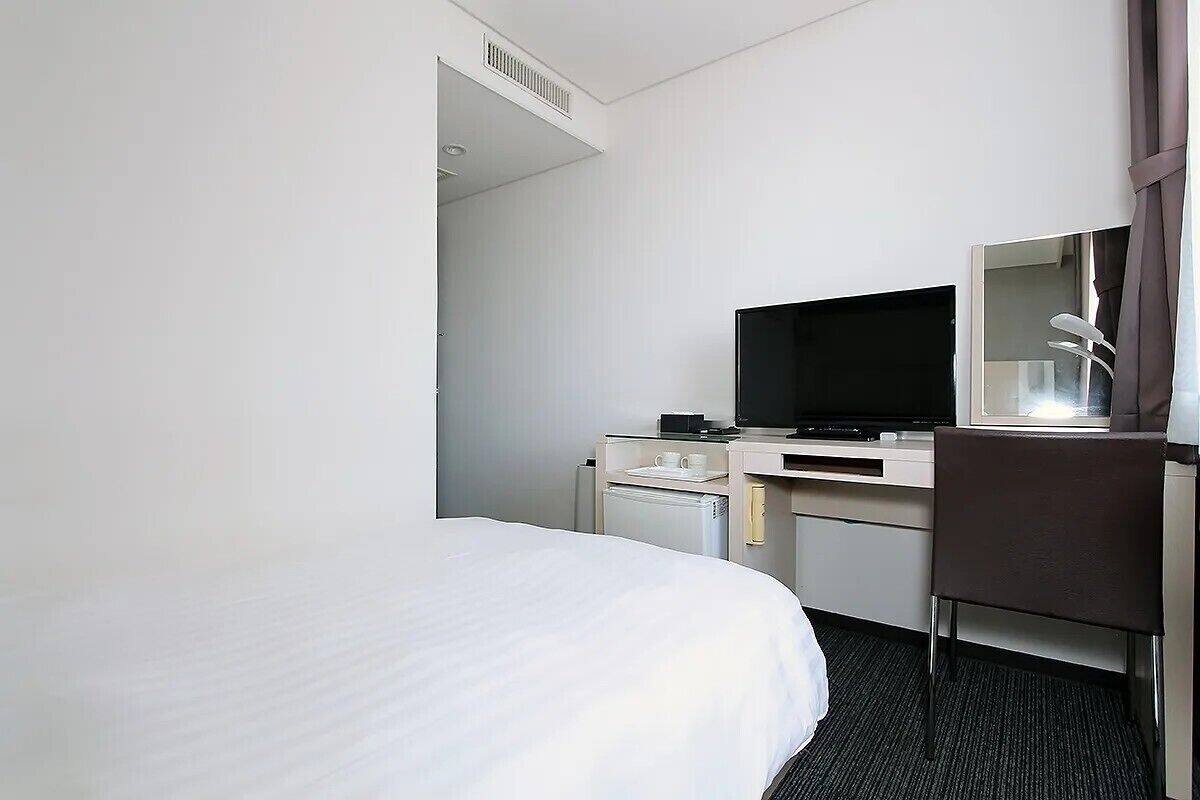 A hotel room with a bed, a desk with a television, a chair, and a small refrigerator.
