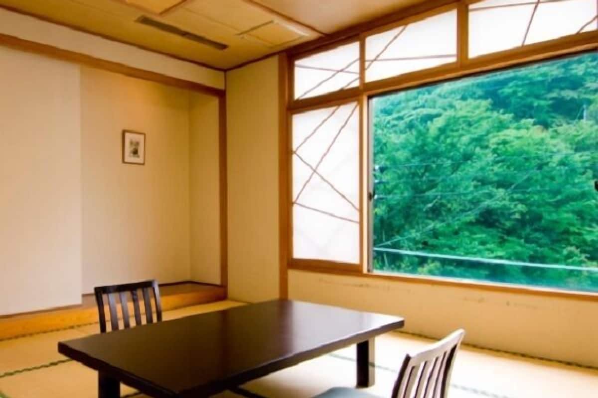 Ein traditioneller japanischer Tatami-Raum mit niedrigem Tisch, zwei Stühlen und einem großen Fenster mit Blick auf Grünflächen.