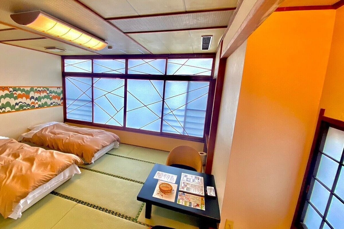 Ein traditioneller japanischer Raum mit Tatami-Böden, zwei Betten mit Futon-Schlafgelegenheiten, einem Fenster mit Shoji-Bildschirmen und einem kleinen Tisch mit einer Speisekarte.