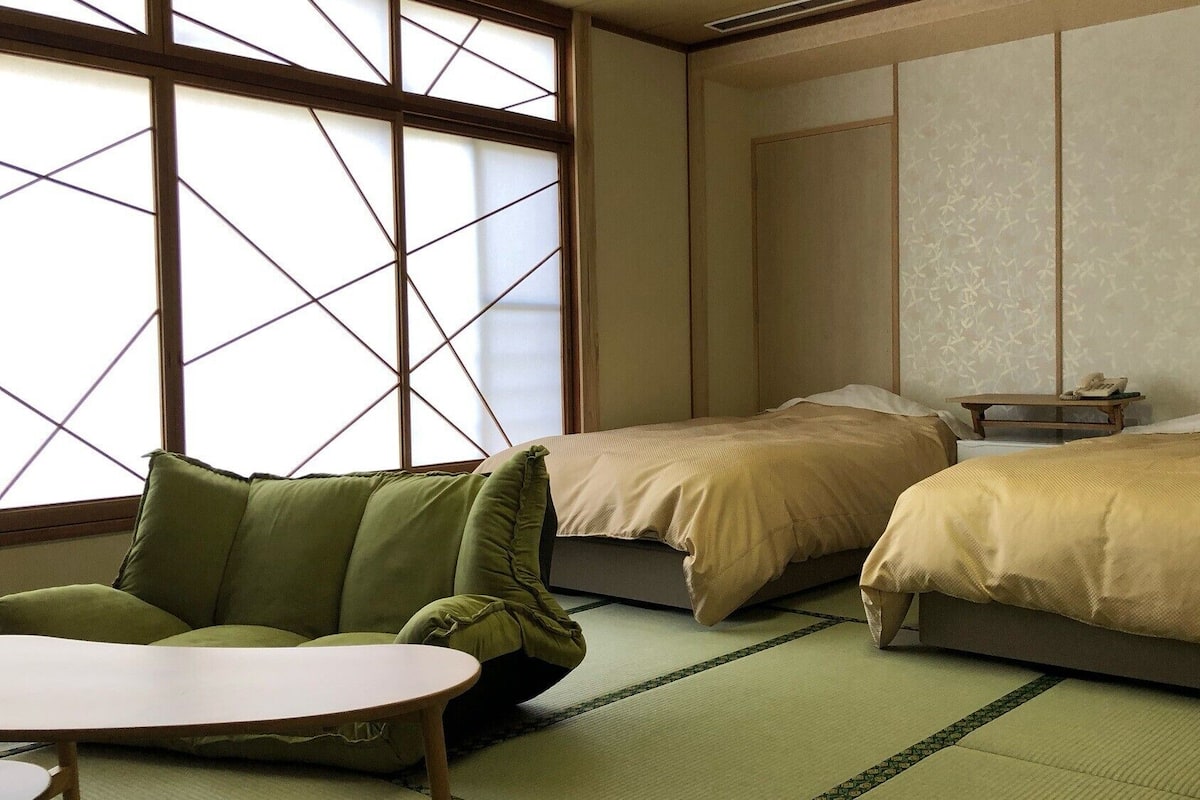 Ein traditionell gestalteter japanischer Raum mit Tatami-Böden, zwei Betten, einem niedrigen Tisch und einem grünen Sofa.