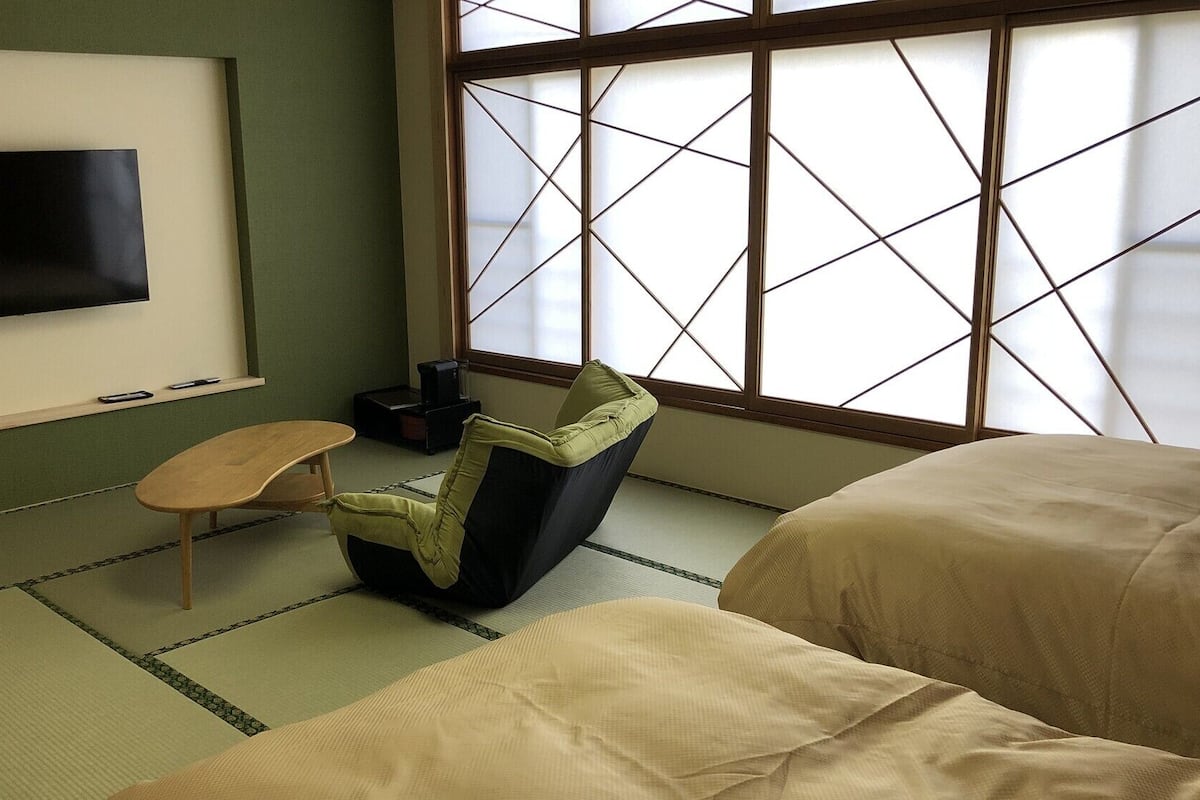 Ein Zimmer mit Tatami-Boden, einem niedrigen Tisch, einem Stuhl, einem Bett, einem Fernseher und einem großen Fenster mit Gittermuster.