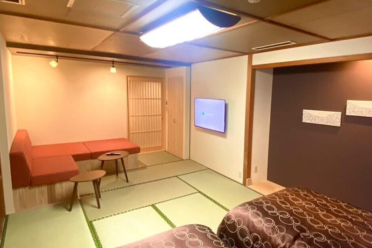 Ein Zimmer mit Tatami-Boden, einem Futon-Bett, einem niedrigen Tisch mit Stuhl, einem Fernseher und einer Schiebetür.