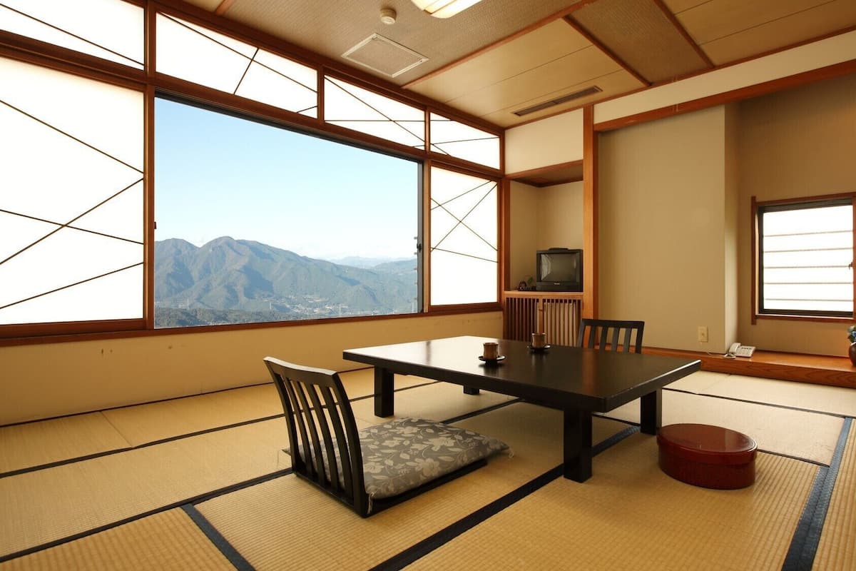 Ein Raum mit Tatami-Matten, einem niedrigen Tisch, Stühlen, einem Fernseher und Blick auf Berge.