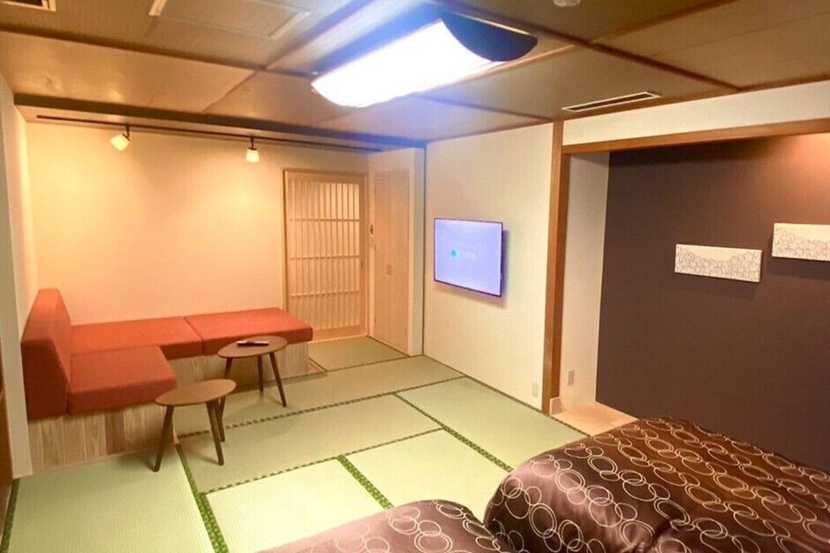 Ein Zimmer mit Tatami-Boden, einem Futon, einem niedrigen Tisch mit Stuhl, einem Fernseher und einer Schiebetür.