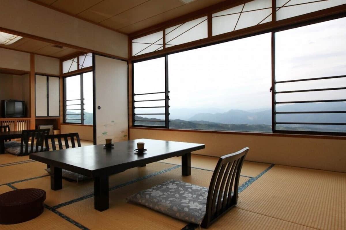 Ein traditioneller japanischer Raum mit Tatami-Boden, einem niedrigen Tisch, Stühlen, einem Fernseher und großen Fenstern mit Blick auf die Berge.