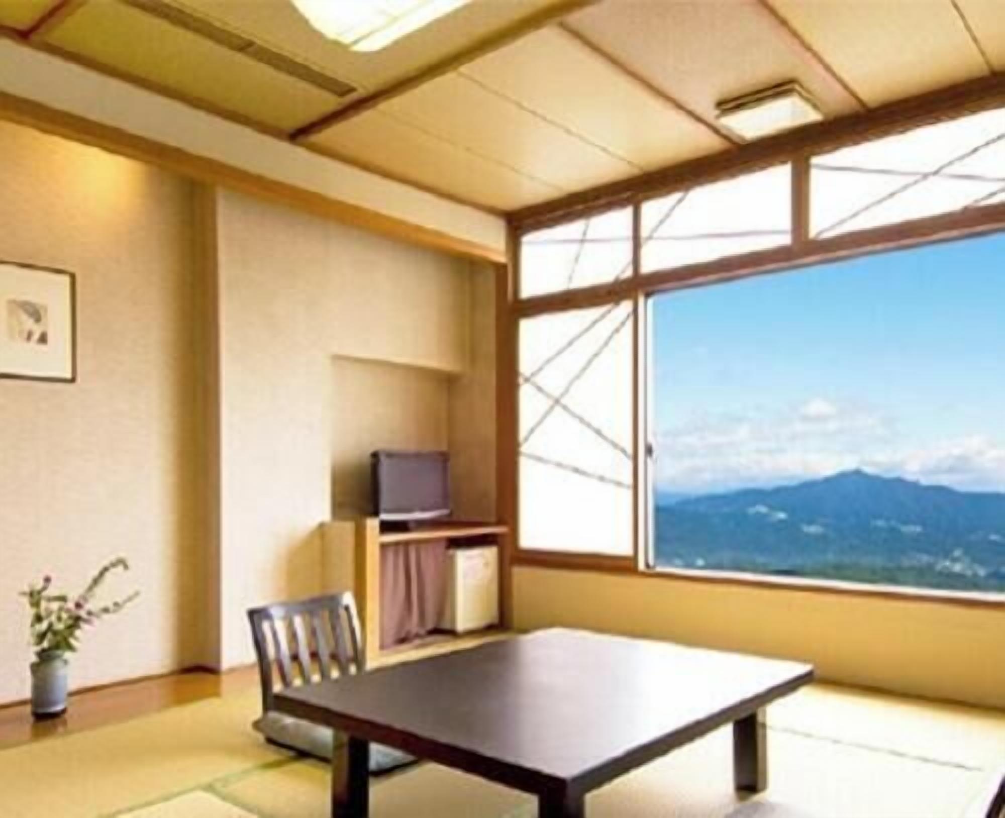 A room with a large window, a table, a chair, a TV, and a small plant.