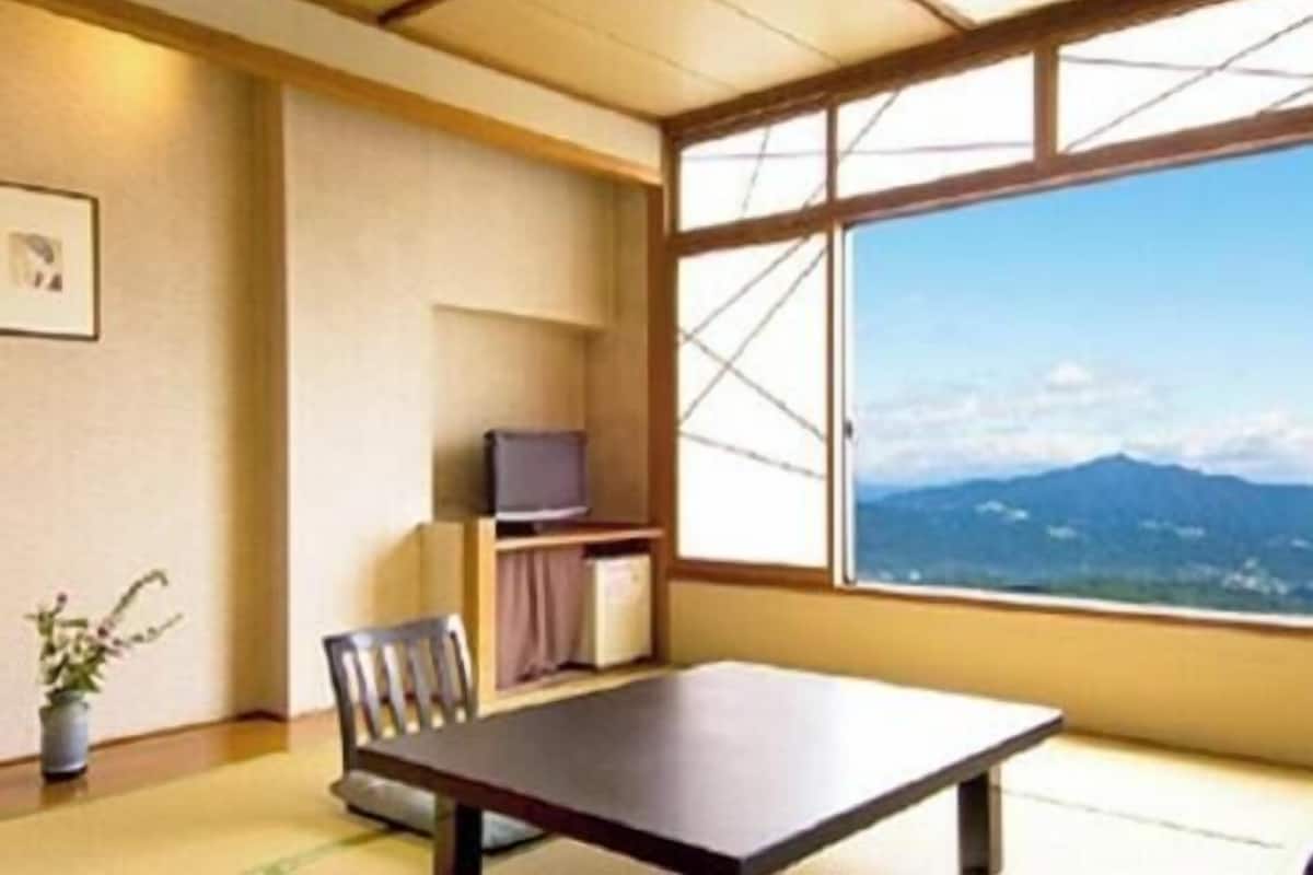 Ein Zimmer mit großem Fenster, Schreibtisch, Stuhl, Fernseher und einer kleinen Ablagefläche.