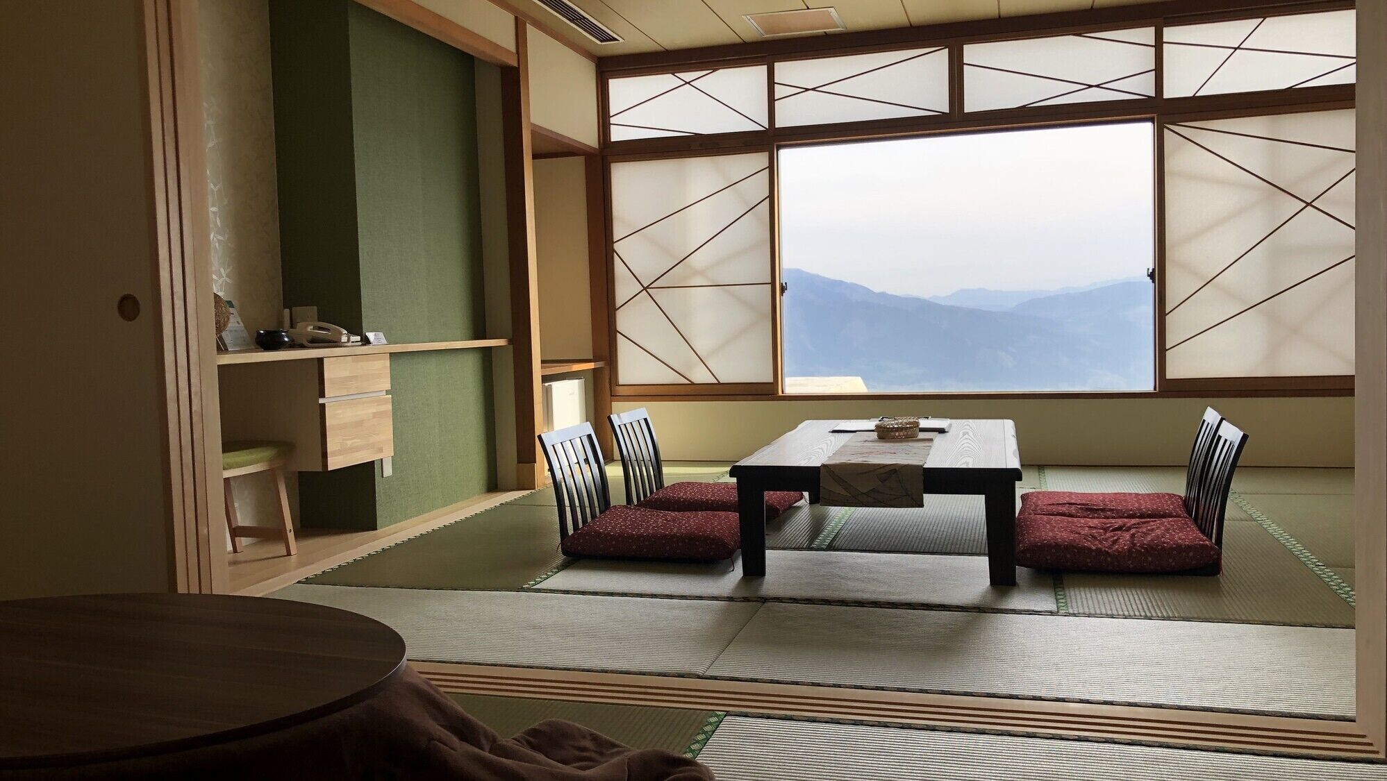 A tatami mat room with a low table and chairs, a sliding shoji screen, and a view of mountains.