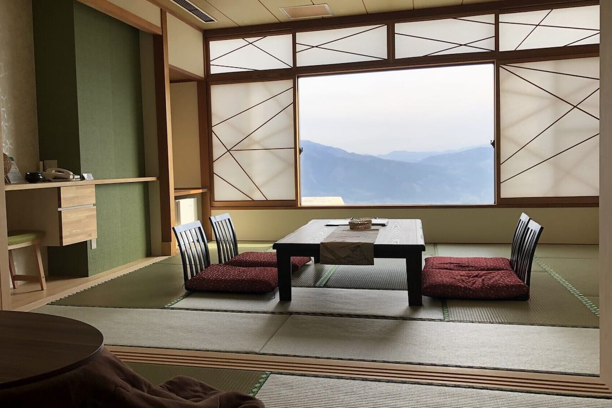 Ein Raum mit Tatami-Matten, einem niedrigen Tisch, Stühlen, einem verschiebbaren Shoji-Bildschirm und Blick auf Berge.