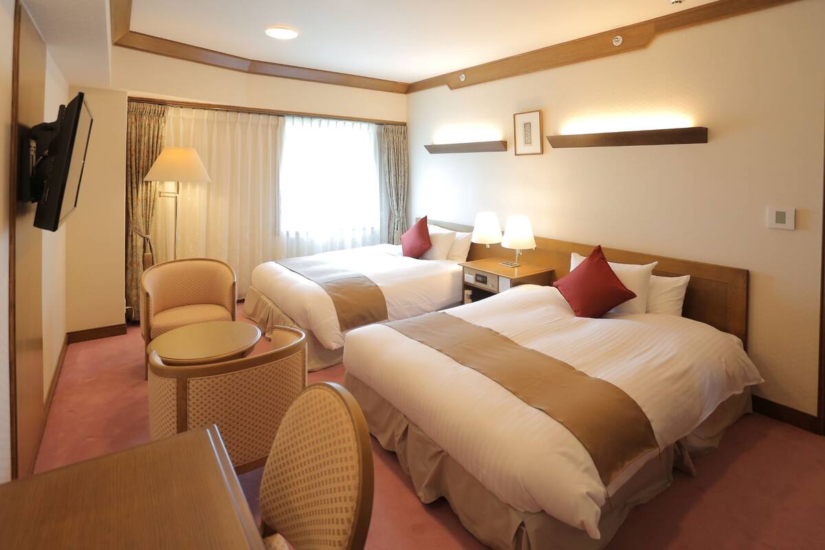 Una habitación de hotel con dos camas, una mesita, una silla, una lámpara y un televisor.