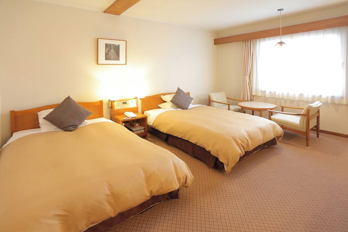 Habitación de hotel con dos camas, una mesita, una silla, una mesita de noche con un teléfono y una ventana con cortinas.
