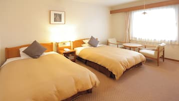 Ein Hotelzimmer mit zwei Betten, einem kleinen Tisch, einem Stuhl, einem Nachttisch mit Telefon und einem Fenster mit Vorhängen.
