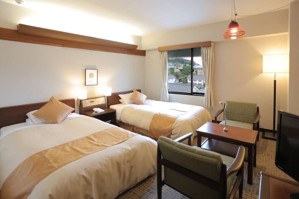 Habitación de hotel con dos camas, un escritorio, una silla, un televisor y una ventana con vistas a edificios.