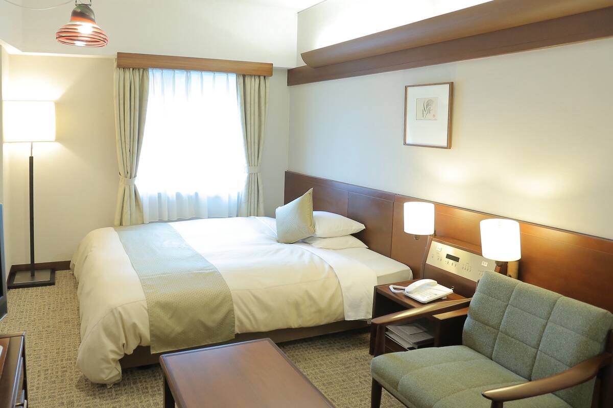 Habitación de hotel con cama, mesita de noche, silla, lámpara y ventana con cortinas.