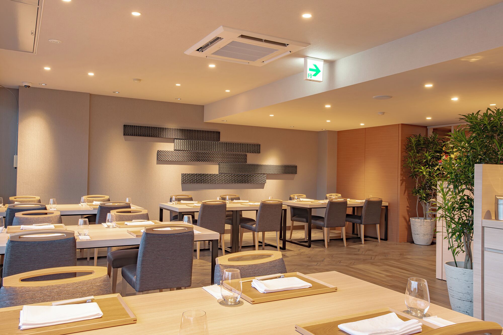 A modern dining area with multiple tables and chairs, a wall-mounted light fixture, and a visible exit sign.