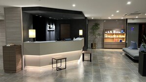 A modern reception area with a reception desk, two lamps, a small table, a potted plant, and a seating area with a sofa and cushions. - Onomichi Royal Hotel (Onomichi)