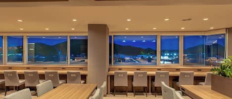 Un comedor moderno con varias mesas y sillas, amplias ventanas que ofrecen vista nocturna de la ciudad.