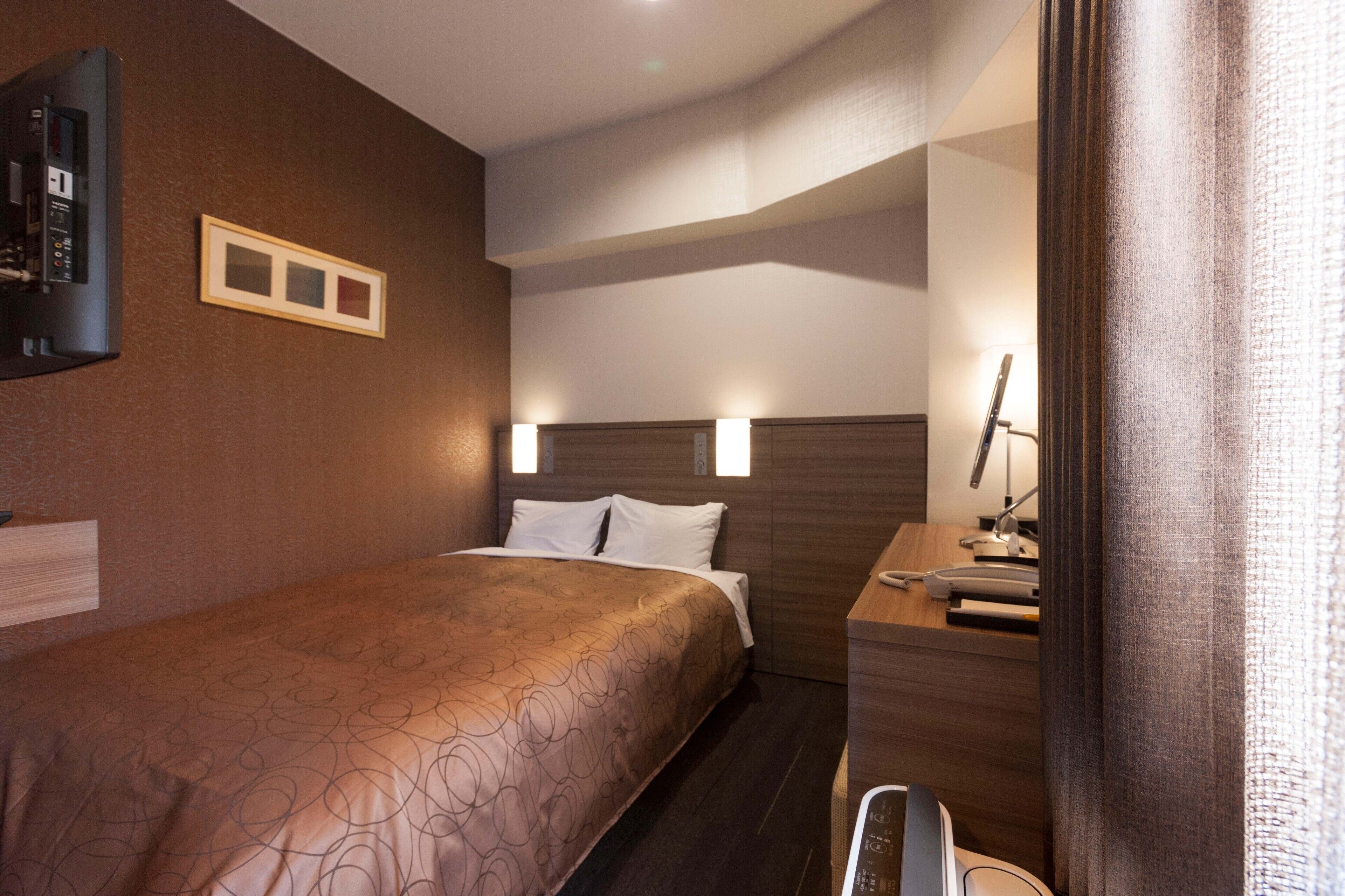 Una habitación de hotel con cama, mesita de noche, lámpara y un escritorio pequeño con una silla.