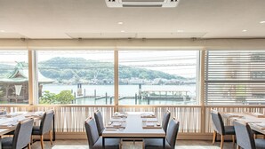 A restaurant with a view of a river and hills, featuring wooden tables and chairs, and large windows. - Onomichi Royal Hotel (Onomichi)