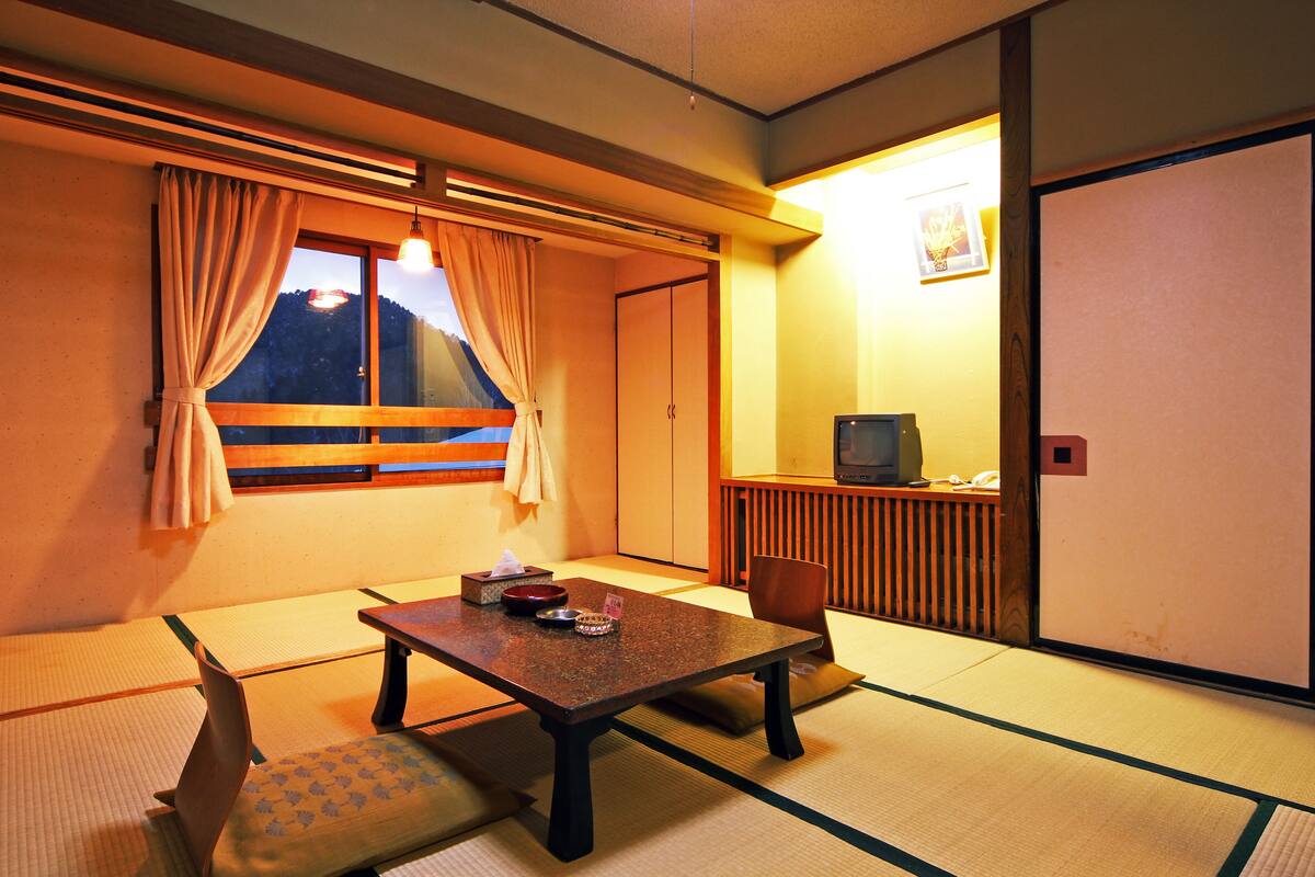 A traditional Japanese room with tatami flooring, a low table, a cushioned bench, a television, and a sliding door.