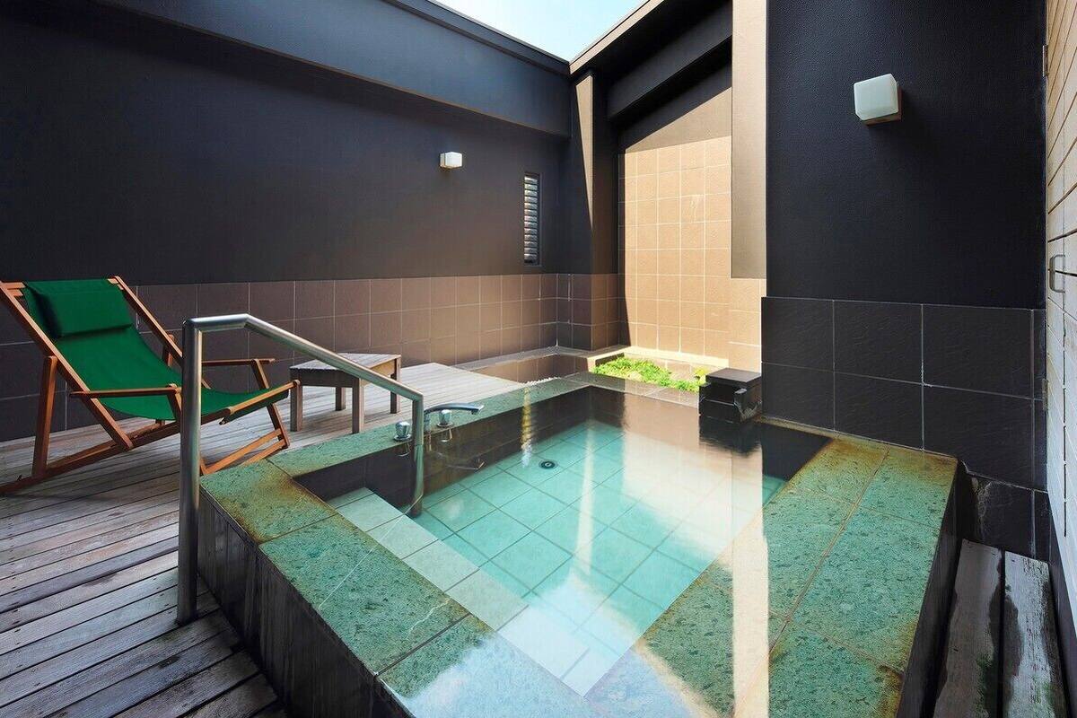 Uma área de spa moderna com um deck de madeira, uma cadeira verde, uma piscina pequena e paredes escuras.