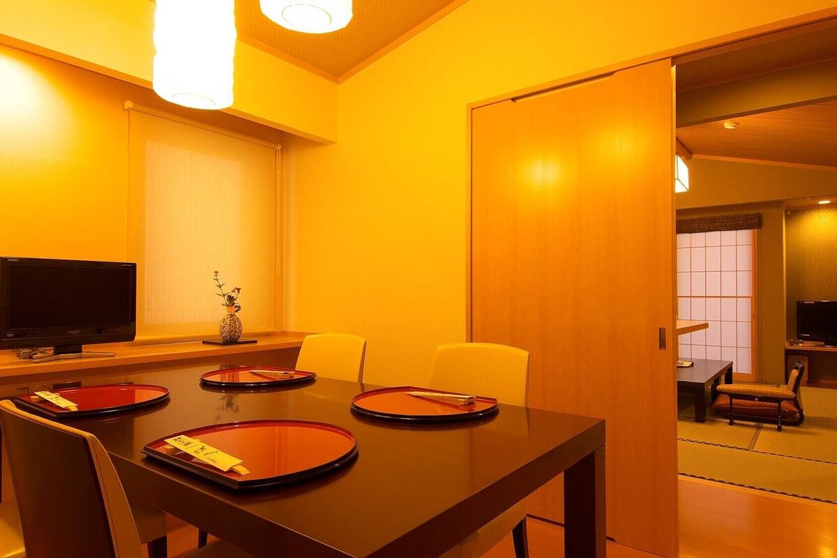 Quarto de hotel com área de refeições, incluindo uma mesa de madeira, cadeiras e uma televisão.