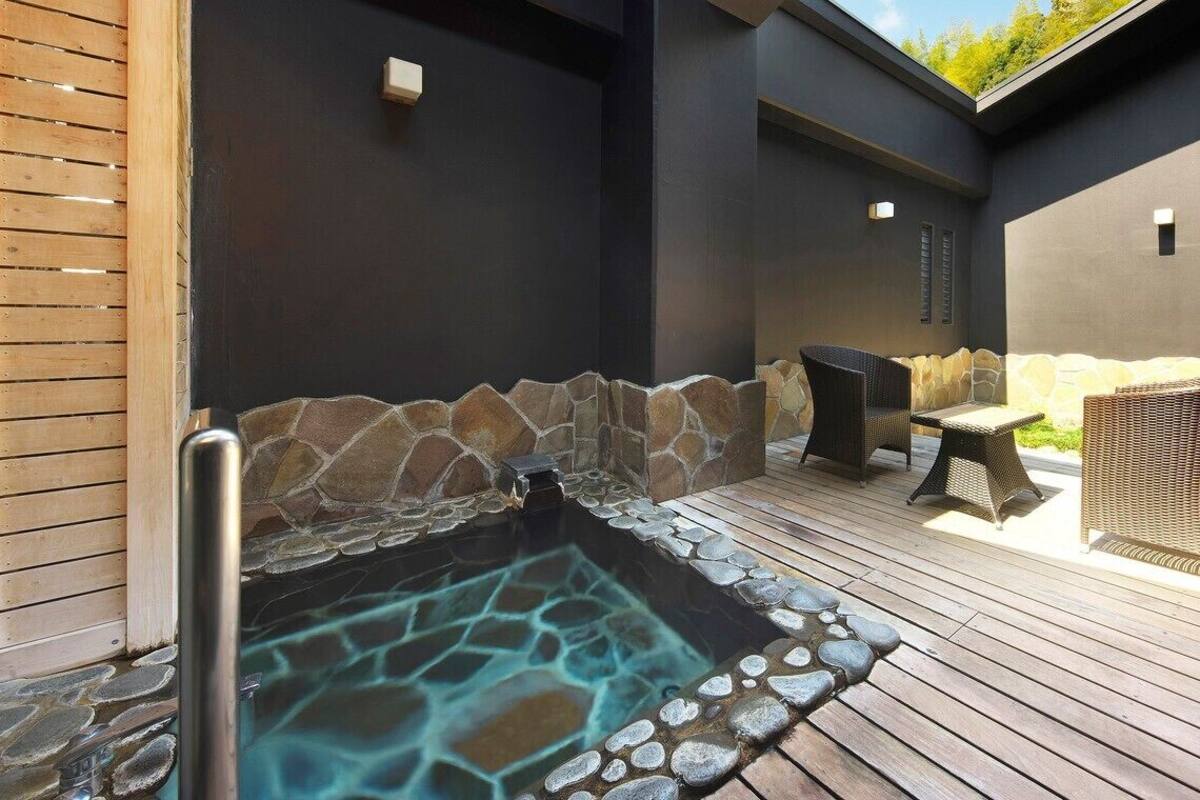 Banheira de hidromassagem com paredes de pedra e deck de madeira, cercada por móveis de vime e um muro de pedra.