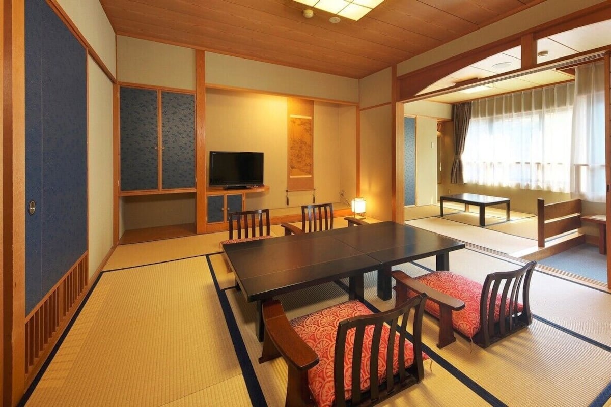 Um quarto japonês tradicional com piso de tatame, uma mesa baixa, cadeiras, uma televisão e portas deslizantes.