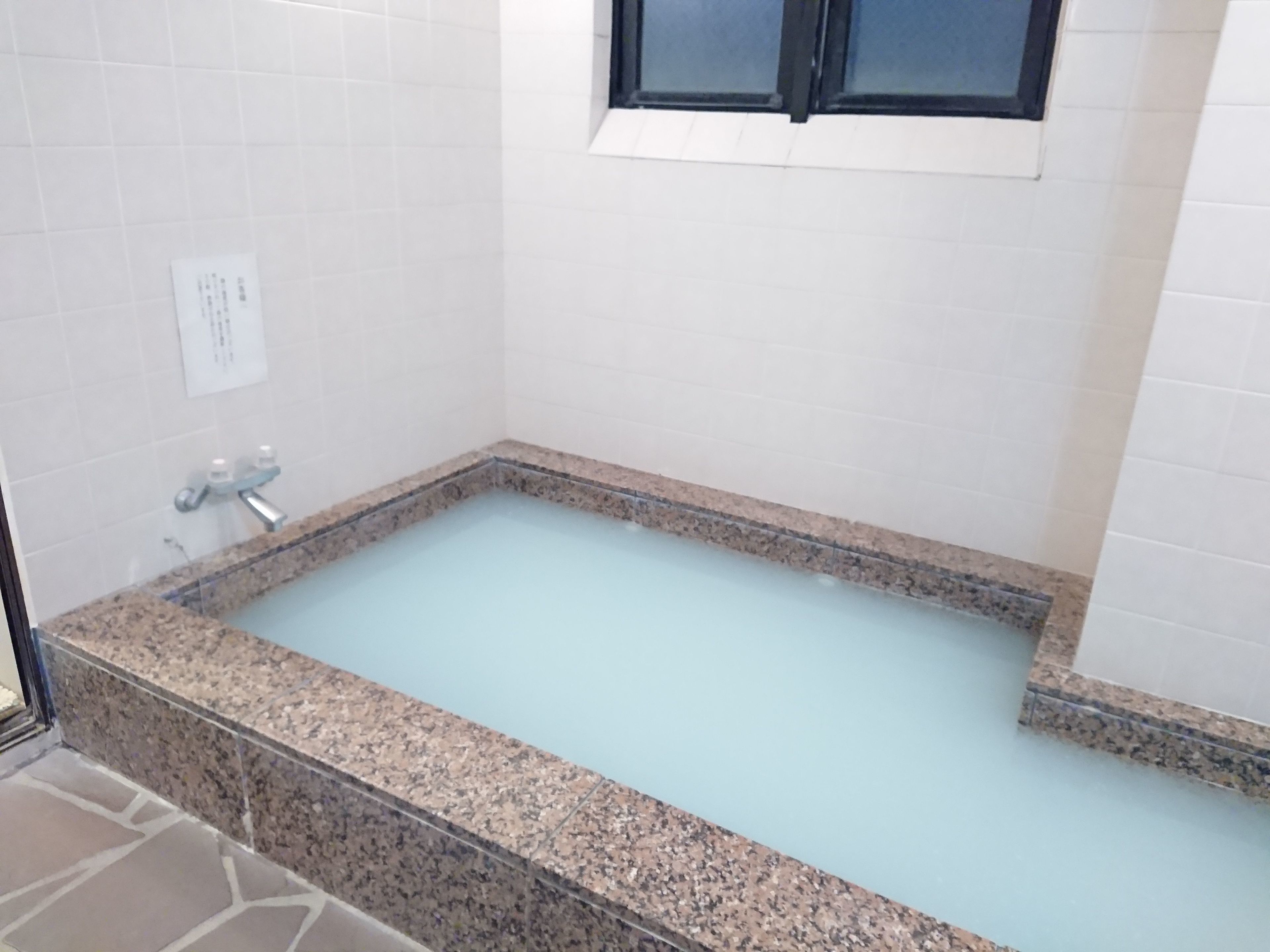 Uma banheira retangular com borda de granito, uma janela acima e paredes revestidas de azulejos brancos.