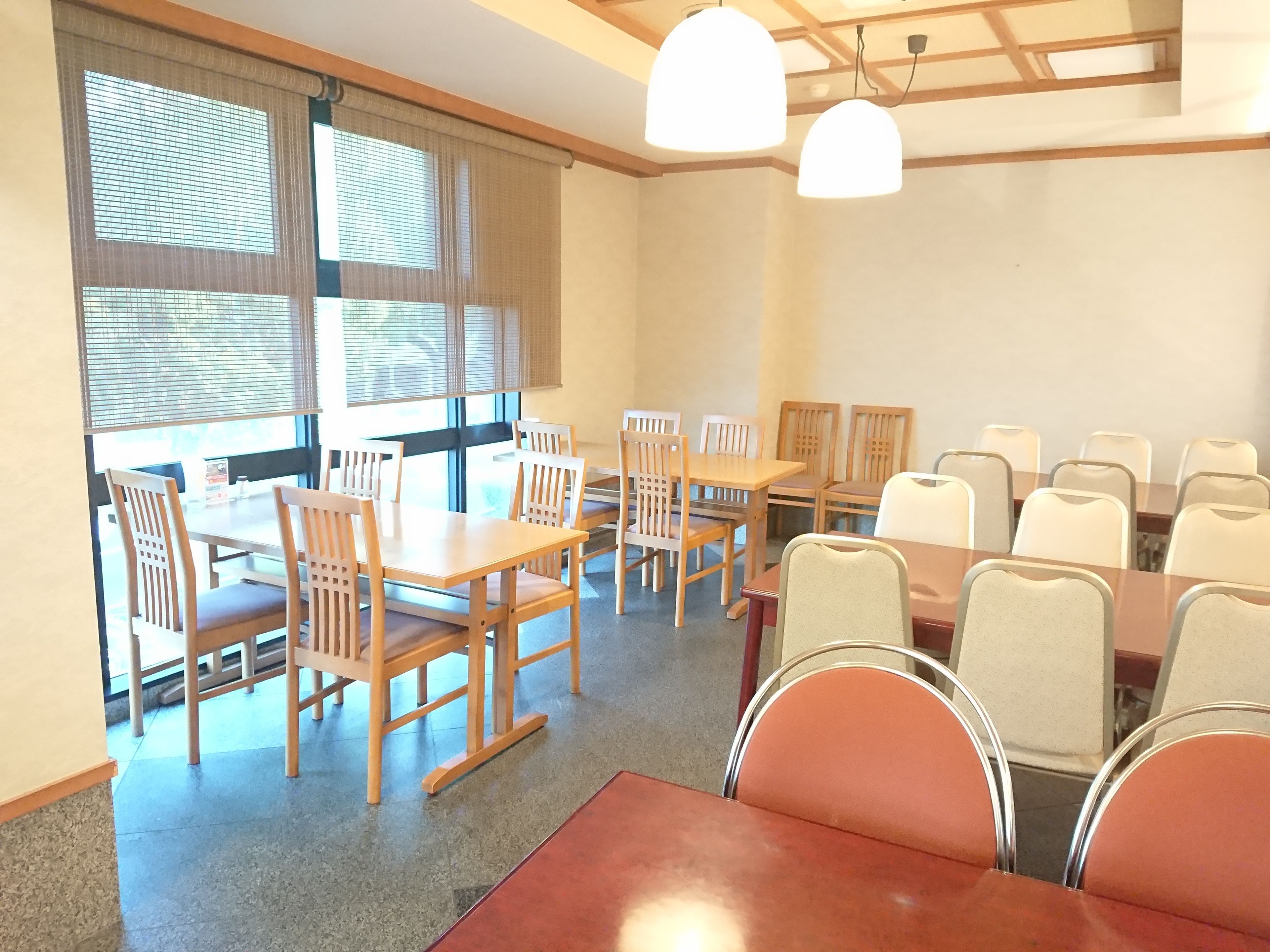 Un comedor con mesas y sillas de madera, ventanas amplias con persianas y lámparas colgantes.