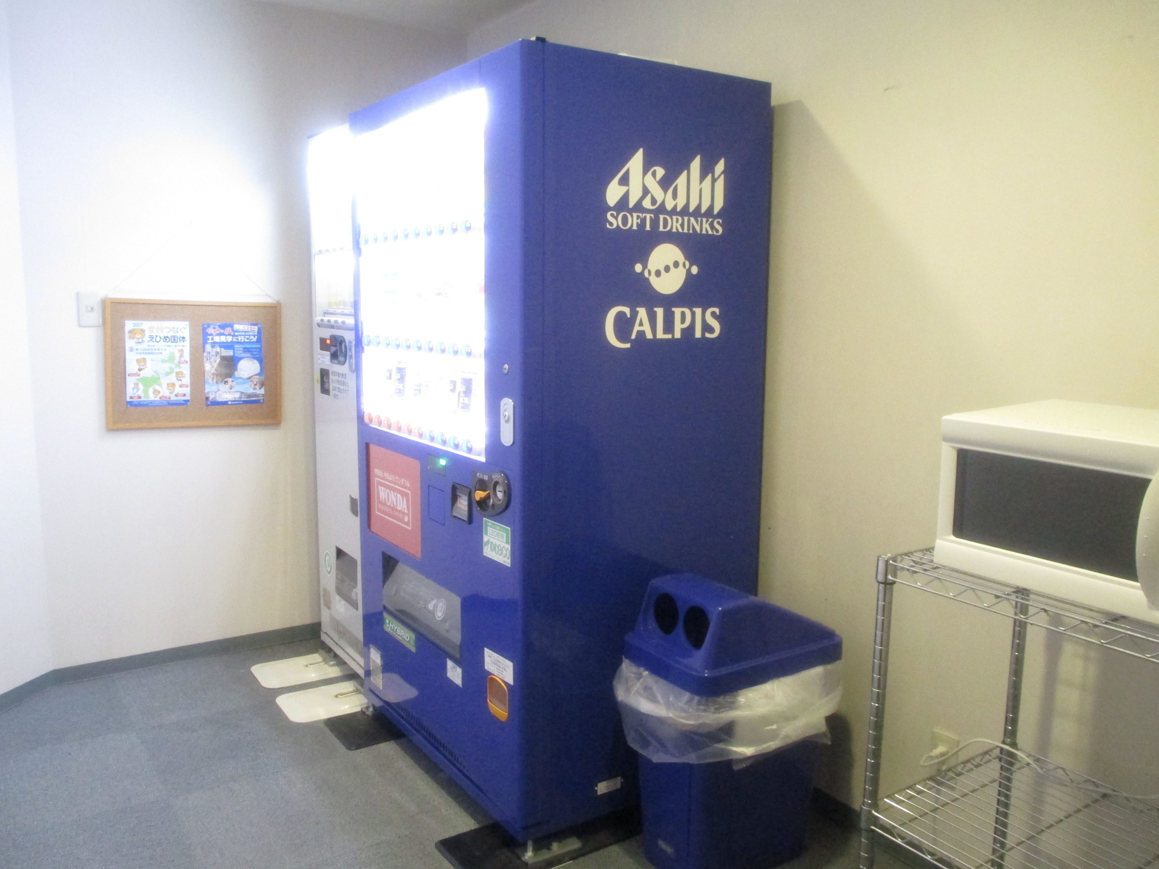 A vending machine labeled "Asahi Soft Drinks" and "Calpis" in a room with a trash bin and a microwave on a shelf.