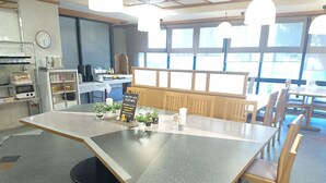 Un restaurante con una larga mesa, sillas y plantas en macetas. Hay un reloj en la pared y un letrero con texto en japonés.
