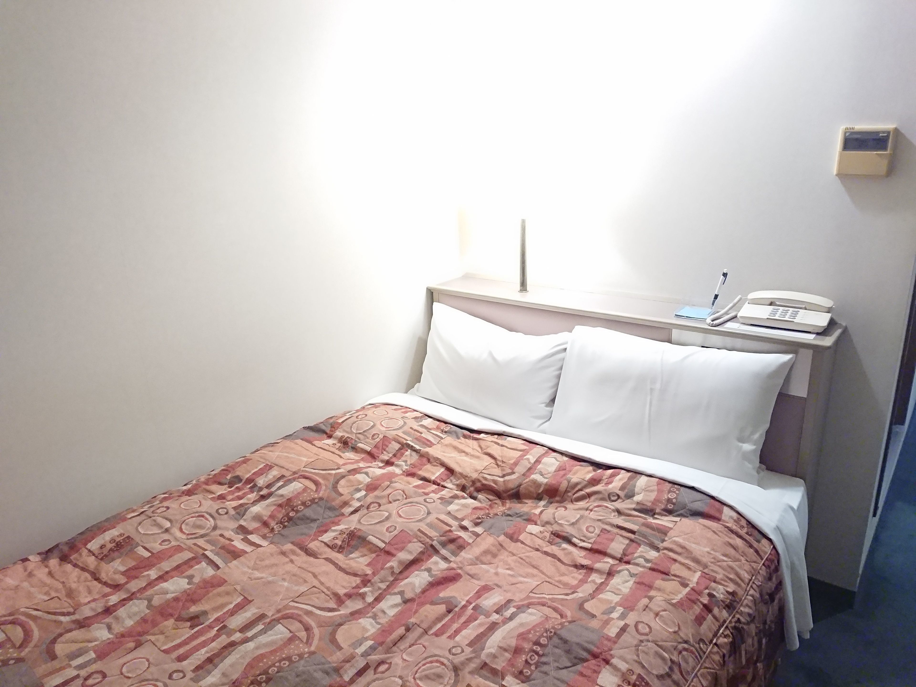 A hotel room with a bed, white pillows, a patterned bedspread, a bedside telephone, and a small wall-mounted device.