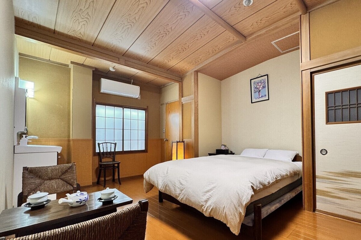 Una habitación tradicional al estilo japonés con techo de madera, una cama, una mesa de comedor con vajilla de té y una ventana con vistas.