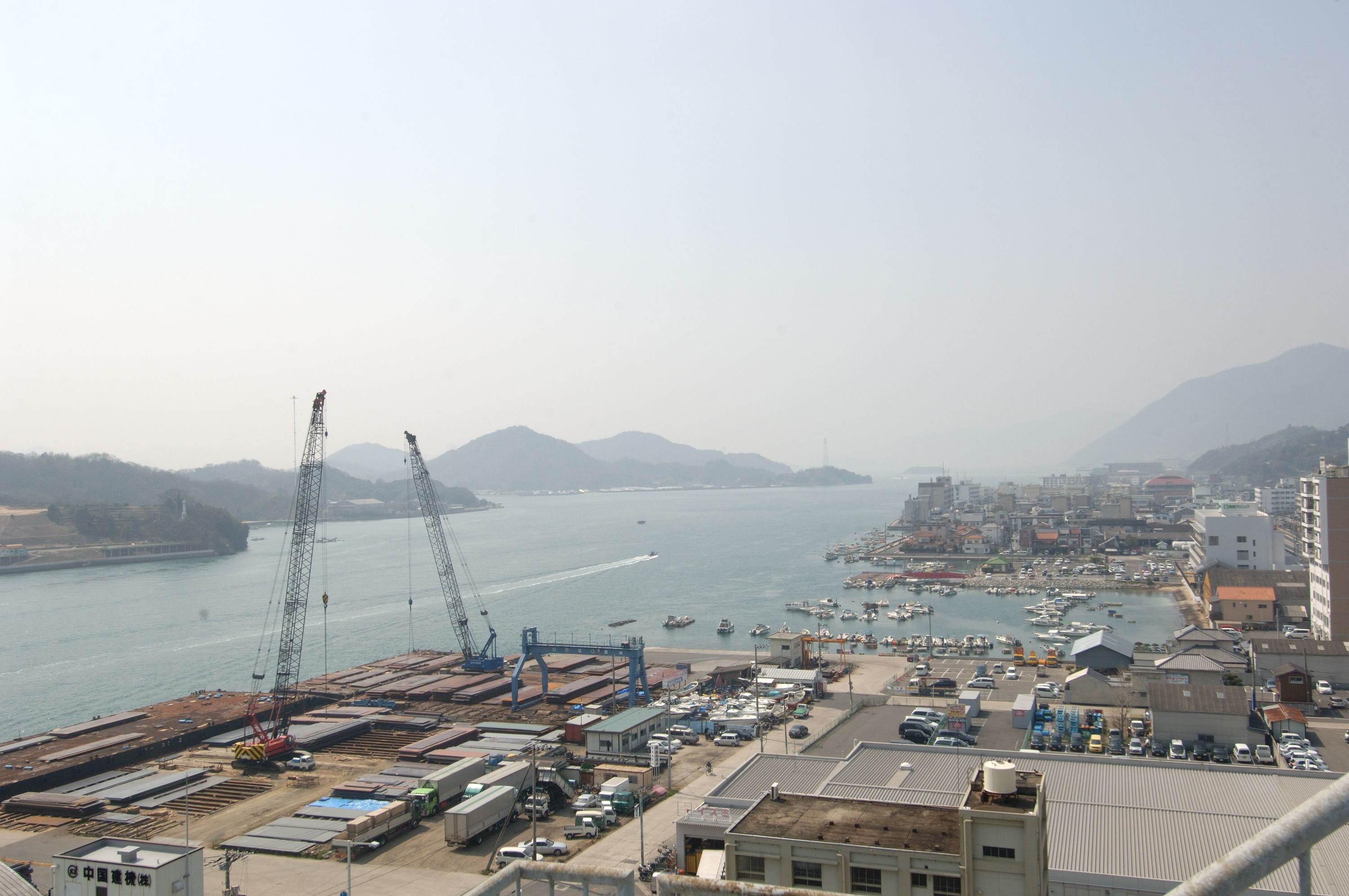 A coastal industrial area with cranes, shipping containers, and a marina with boats.