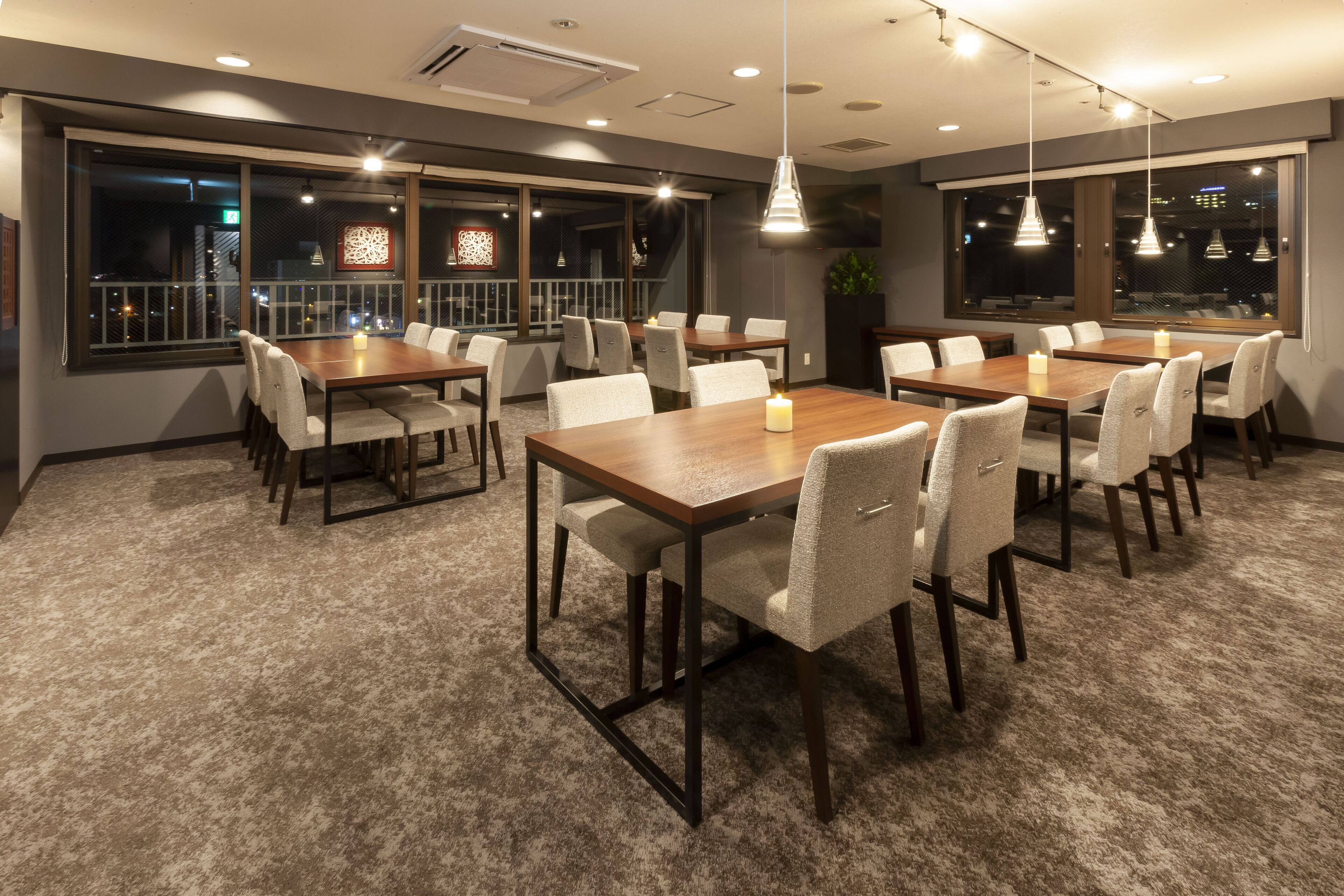 A conference room with multiple tables and chairs, large windows with city views, and pendant lighting.