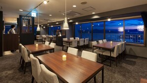 Un comedor de hotel con varias mesas y sillas, un bar y amplios ventanales que ofrecen vistas de la ciudad de noche.