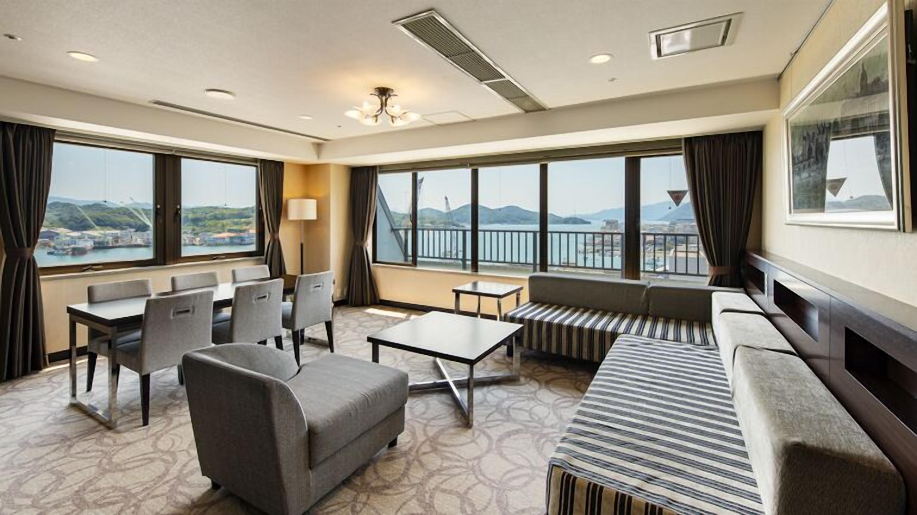 Habitación de hotel con sofá, sillas, una mesa y ventanas amplias que ofrecen vistas al mar y a las colinas.