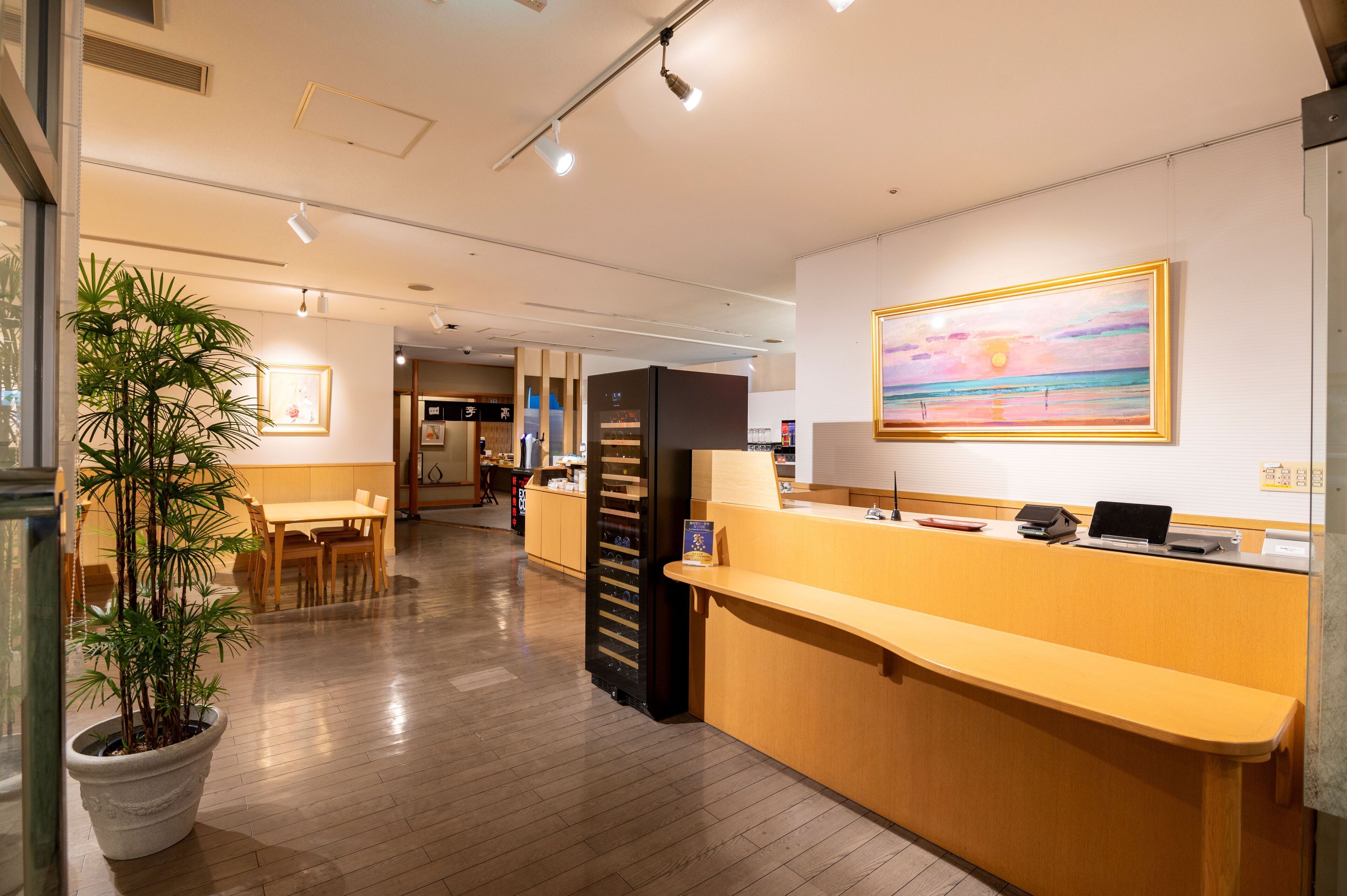 A hotel reception area with a wooden reception desk, a large painting on the wall, and a potted plant.