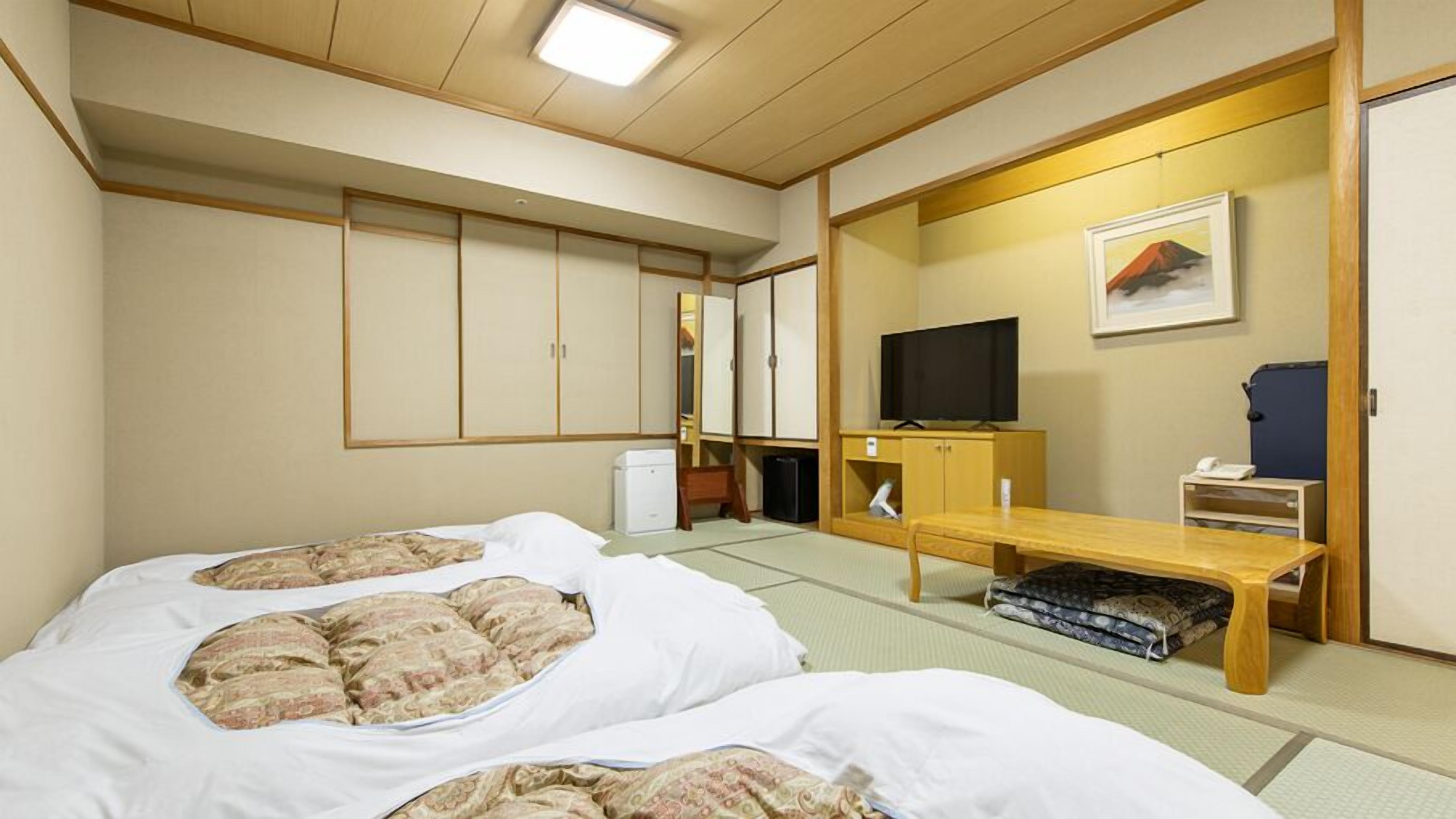 Habitación de hotel tradicional al estilo japonés, con suelo de tatami, una cama con ropa de cama estilo futón, un televisor, un escritorio con silla y un armario con puertas correderas.