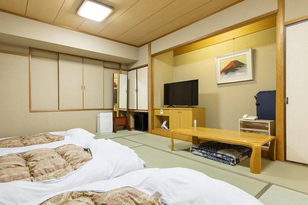Et traditionelt japansk hotelværelse med tatamimåtter, en seng med futon sengetøj, et fjernsyn, et skrivebord med stol og et garderobeskab med skydedøre.